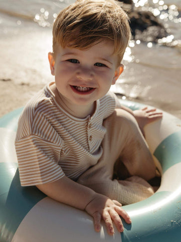 Toddler boy smiling to the camera sitting on a back float wearing a neutral striped baby tee 