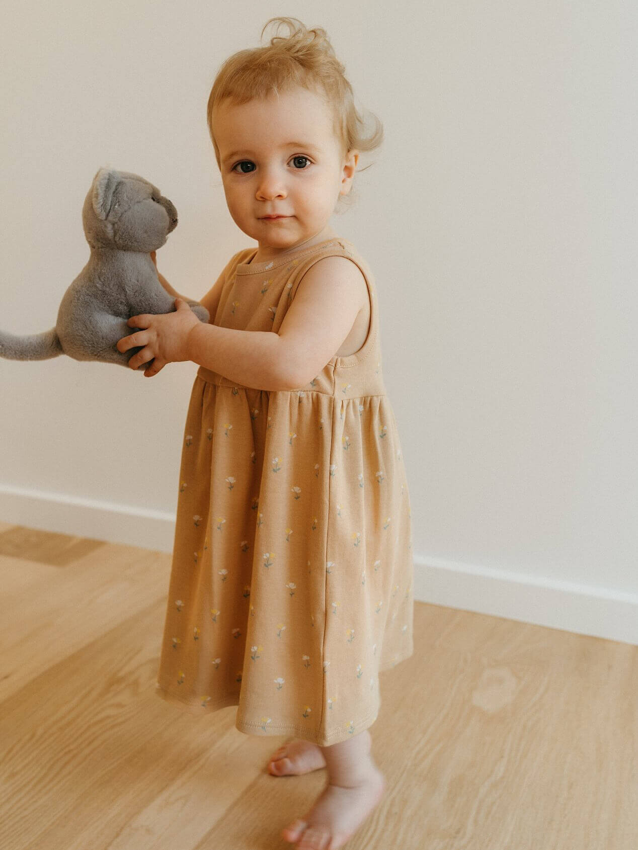 Child in a brown dress holding a gray stuffed animal on a wooden floor.