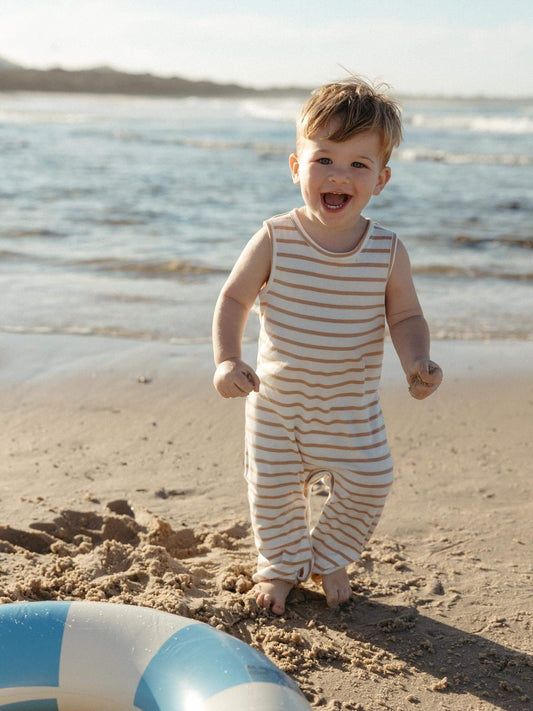 A toddler stands barefoot on a sandy beach wearing a sleeveless, one-piece cream and tan striped romper. The ocean waves are visible in the background, and a blue and white striped beach float is partially visible in the foreground.