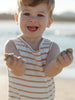 A smiling toddler standing on a beach, wearing a sleeveless white tank top with thin tan horizontal stripes and holding small clumps of sand in both hands.