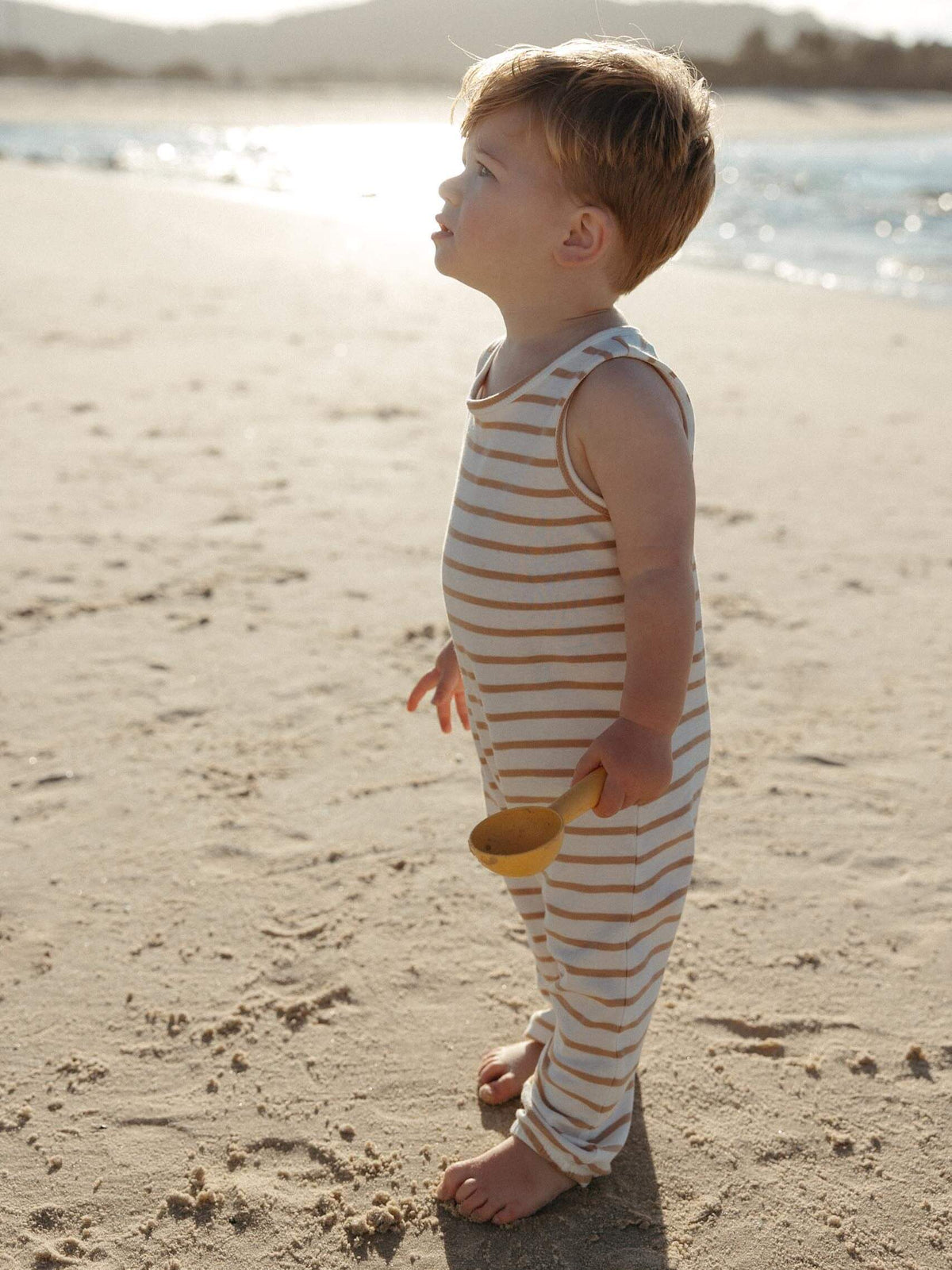 A toddler stands on a sandy beach wearing a sleeveless, one-piece striped romper in beige and white, holding a small yellow shovel.
