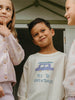 Three children standing together with one wearing a sweatshirt that says 'Yes to Adventures'.