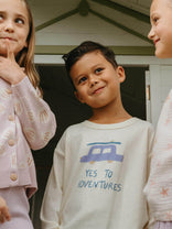 Three children standing together with one wearing a sweatshirt that says 'Yes to Adventures'.