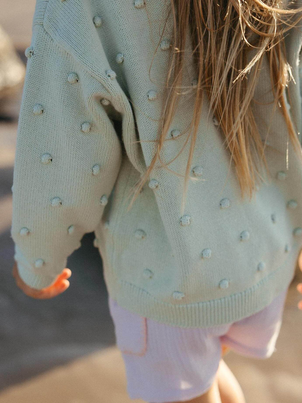 A close-up lifestyle shot of a child wearing a pale mint green knit sweater with raised pom-pom details and light pink shorts, outdoors in warm sunlight.