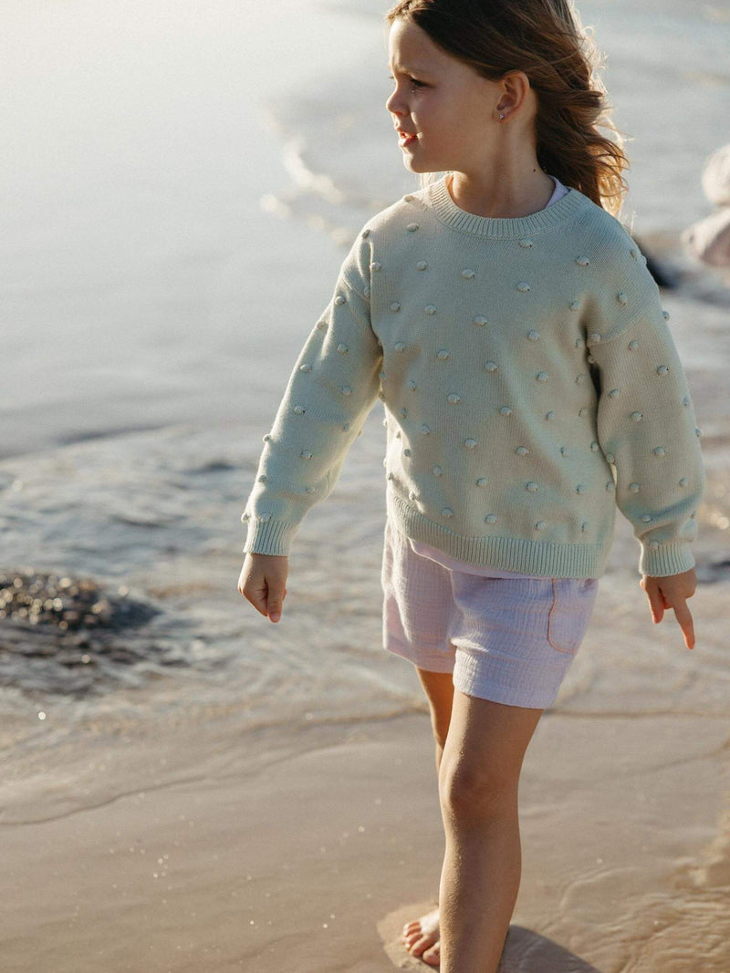 A young girl walking barefoot on a sandy beach near the water, wearing a pale mint green knit sweater with raised pearl-like details and white shorts.