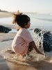 A child with curly hair sits in shallow water on a beach, wrapped in a pale peach towel or wrap, with splashing water around them.