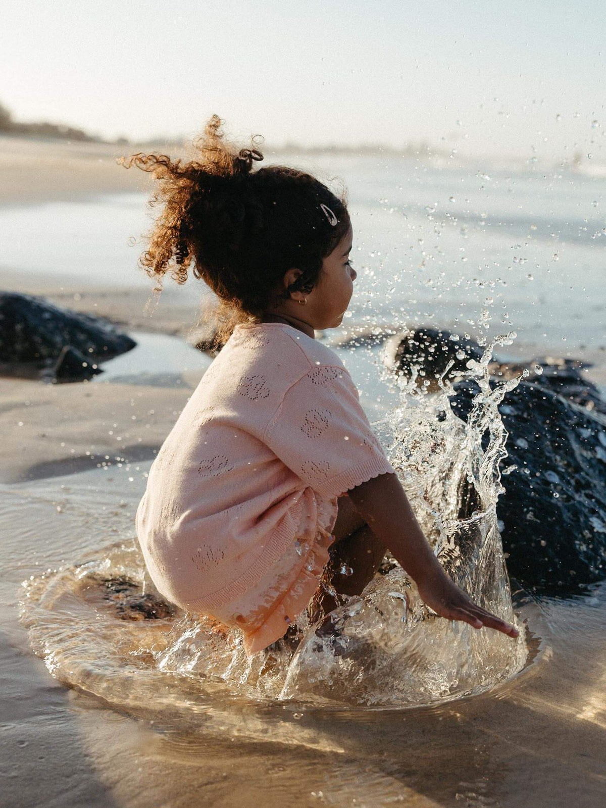 A child with curly hair sits in shallow water on a beach, wrapped in a pale peach towel or wrap, with splashing water around them.