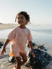 A young child with curly hair stands in shallow water on a beach, wearing a peach-colored short-sleeve t-shirt and matching shorts with a subtle pattern.