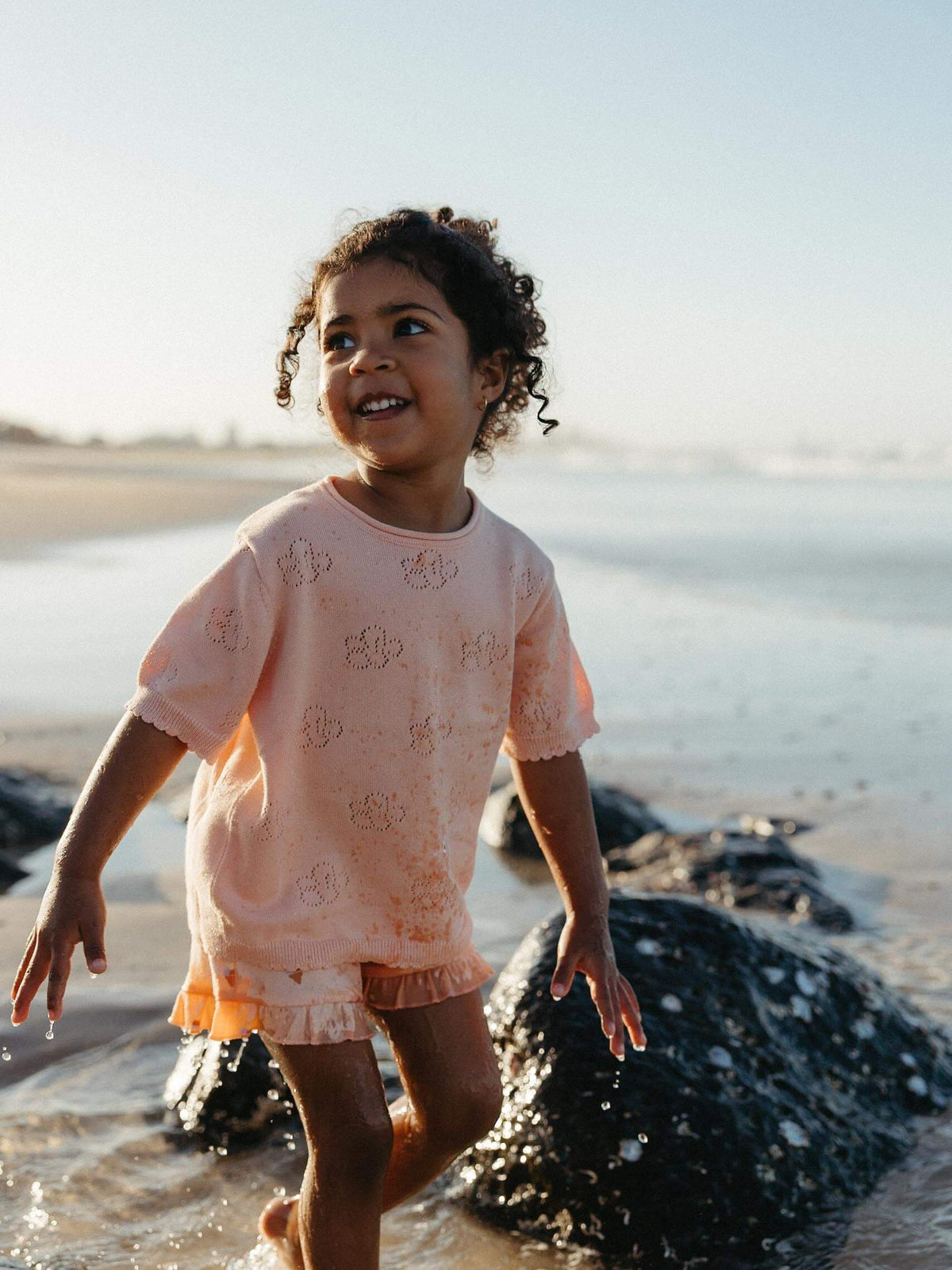 A young child with curly hair stands in shallow water on a beach, wearing a peach-colored short-sleeve t-shirt and matching shorts with a subtle pattern.