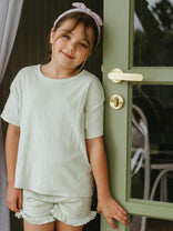 Young girl in a light green outfit standing next to a green door. Lara Towelling Tee Shirt - Mint Girls