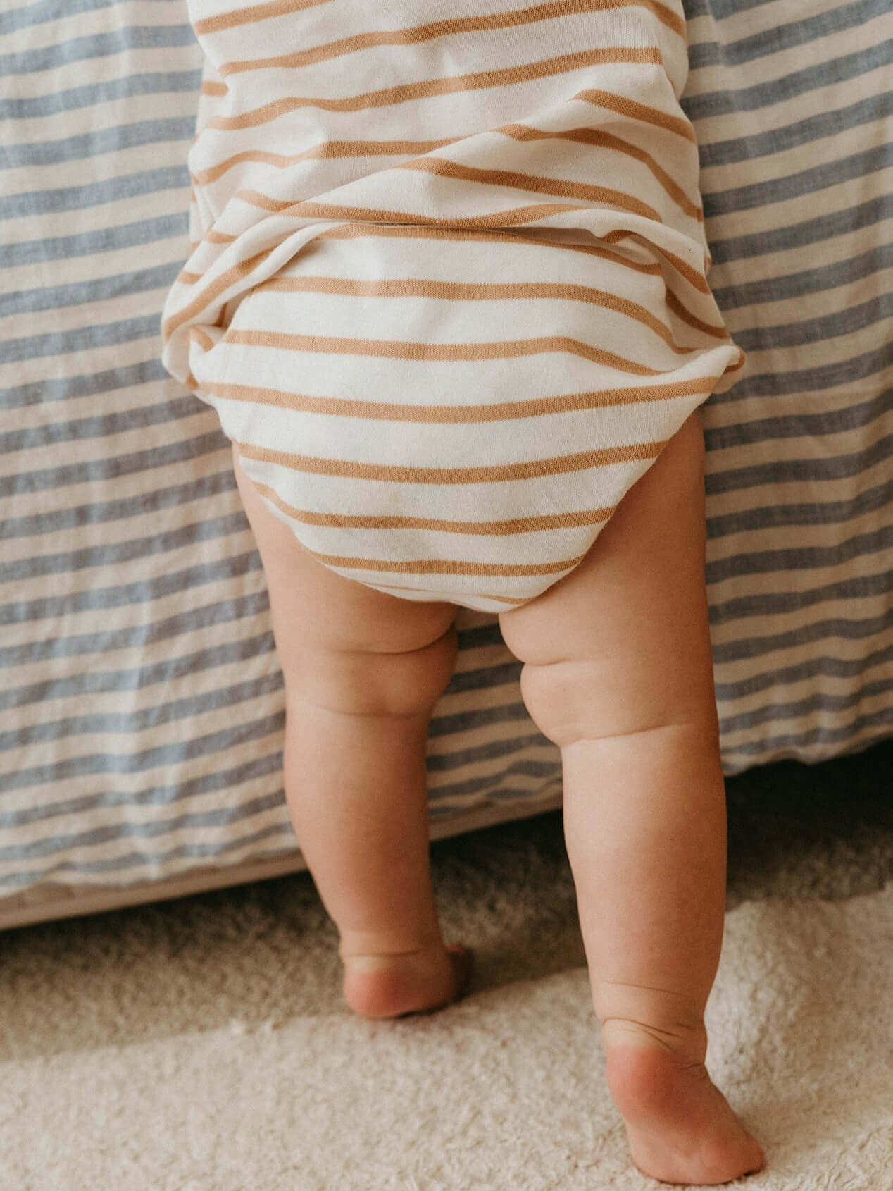 Baby wearing a striped onesie standing on a carpeted floor with a striped couch in the background.