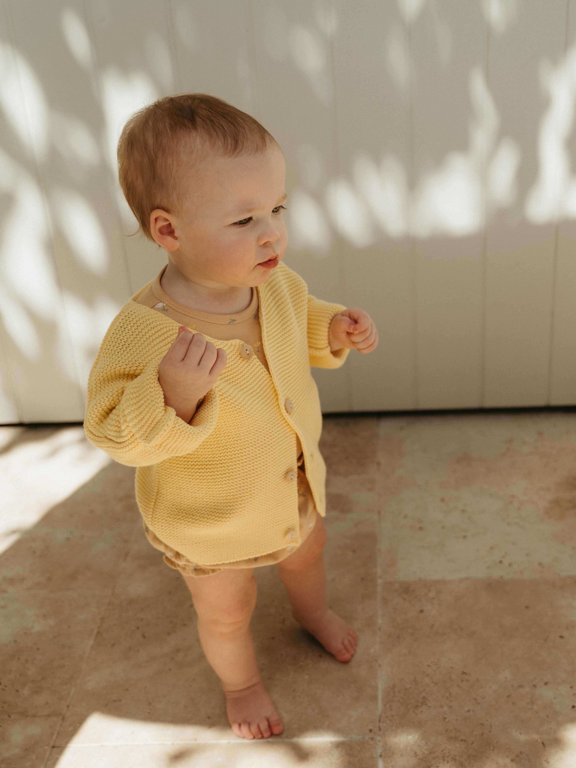 Baby in a yellow outfit standing on a tiled floor with sunlight casting shadows.