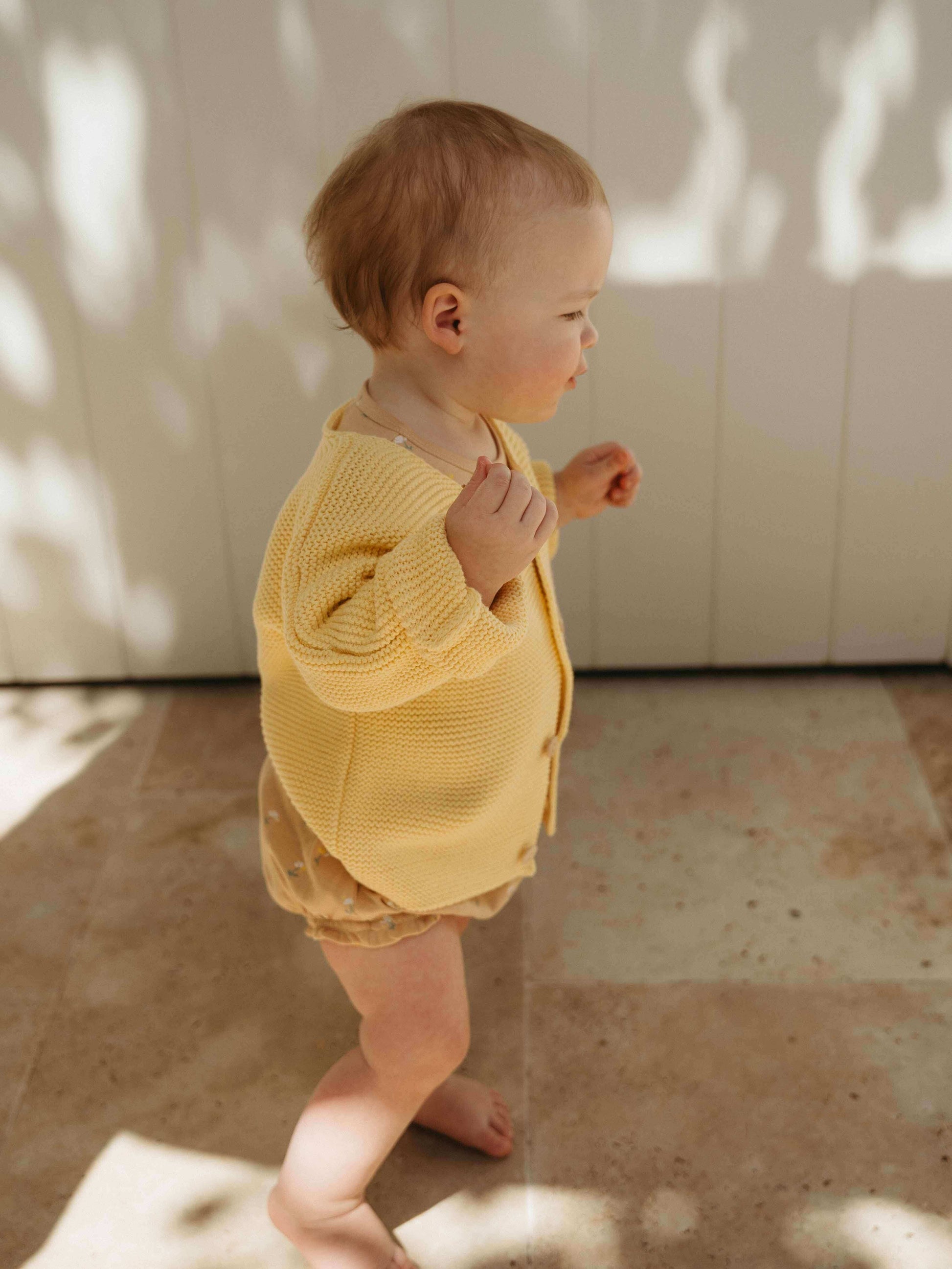Baby in a yellow outfit standing on a tiled floor with sunlight casting shadows.
