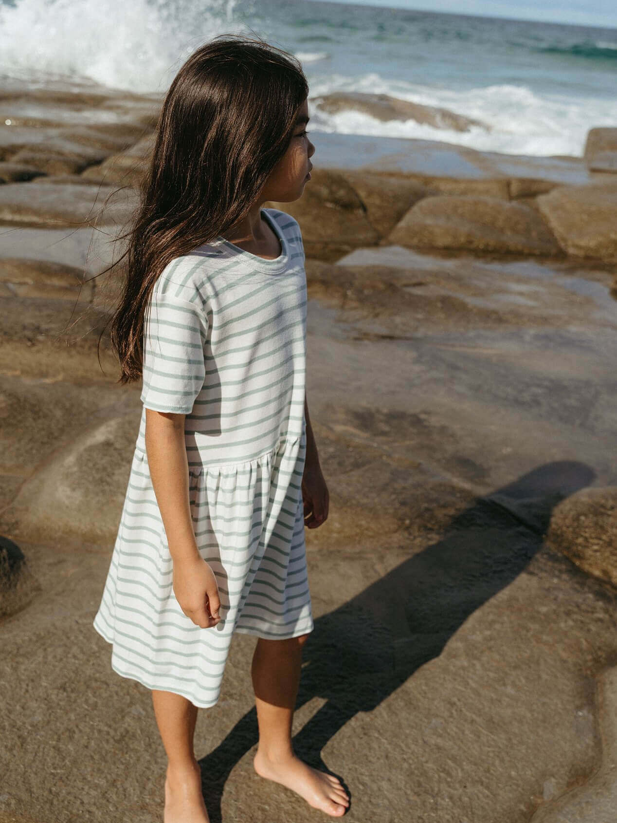 Young girl in a striped dress standing on a rocky beach with ocean waves in the background