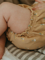 Close-up of a baby wearing a mustard yellow bloomers with floral patterns on a striped fabric background.