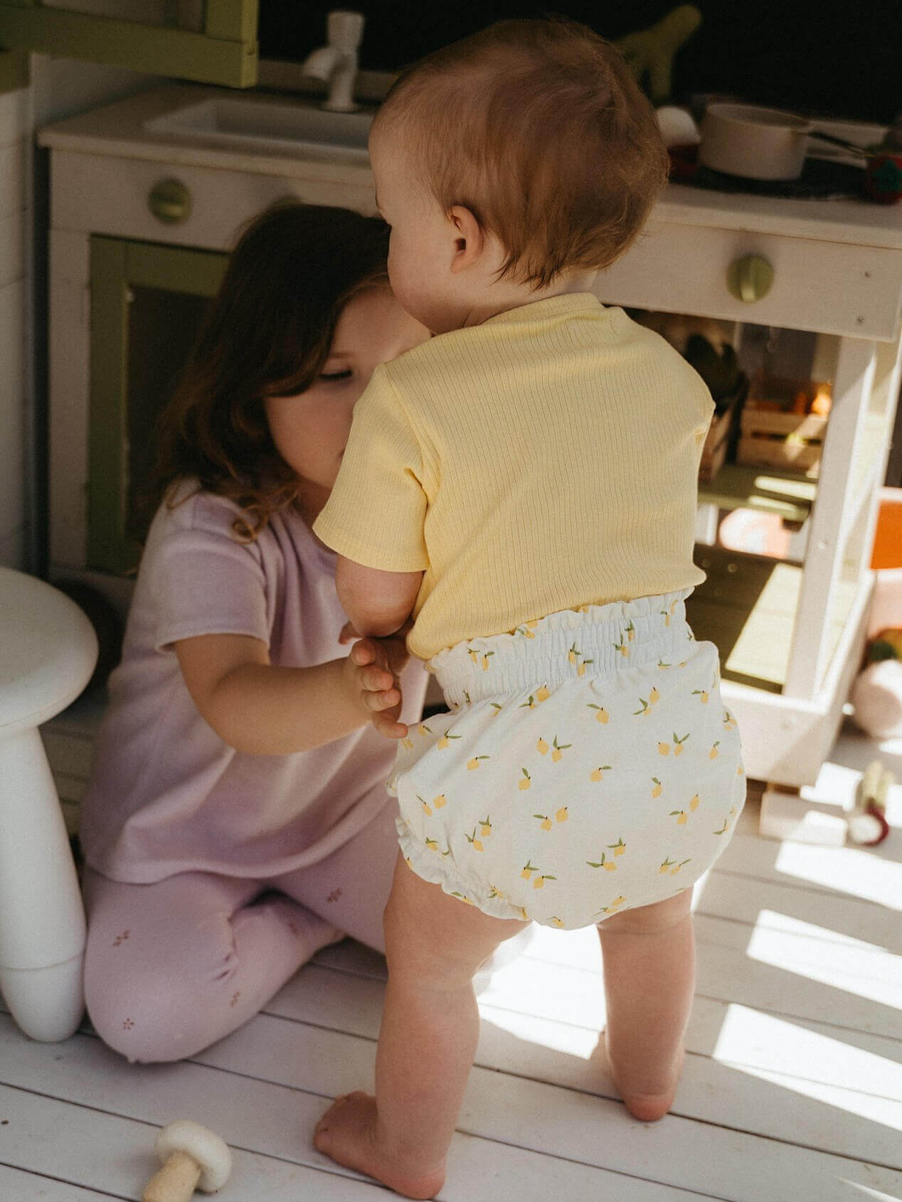 Two children playing together on a wooden floor with a kitchen in the background.