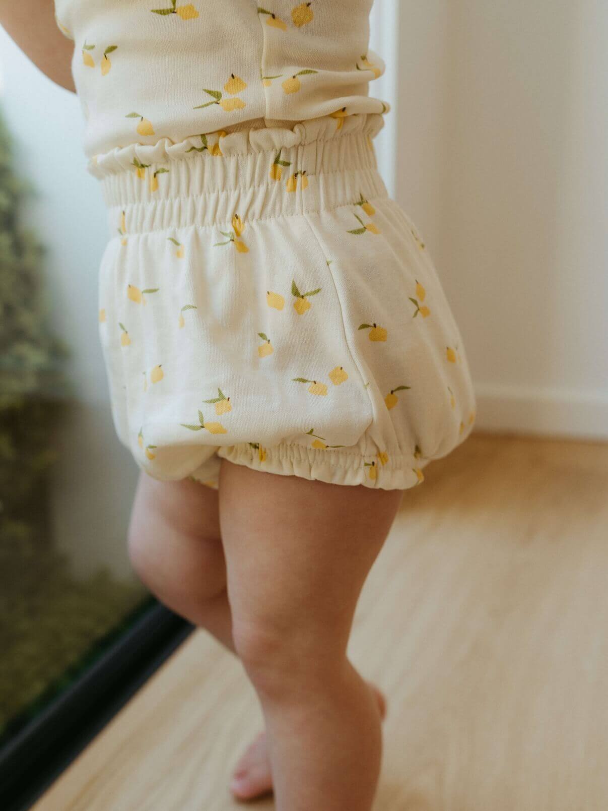 Baby wearing white bloomers with yellow lemon pattern on a light wooden floor.