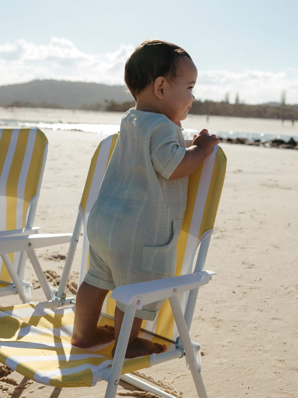 A baby standing on a yellow and white striped beach chair on a sandy beach, wearing a light blue and white textured romper.
