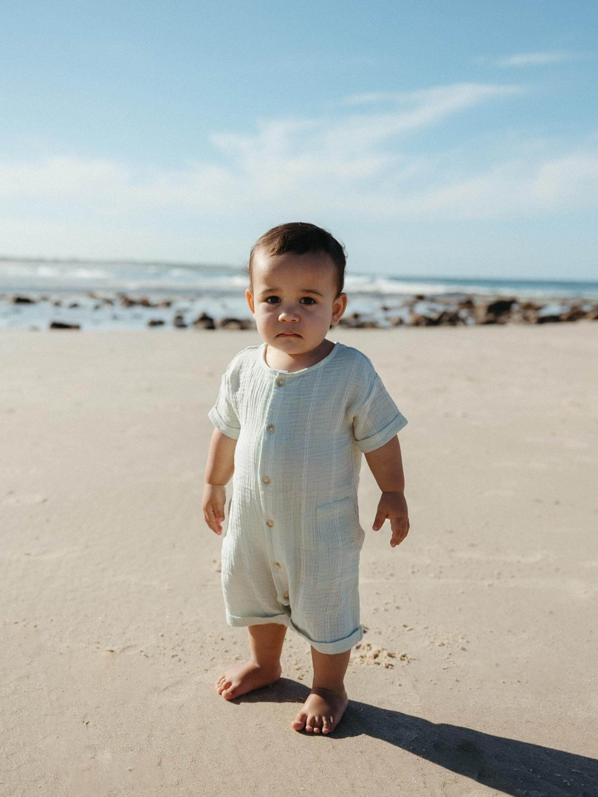 A baby standing on a sandy beach wearing a light cream-colored short-sleeve romper with front buttons.
