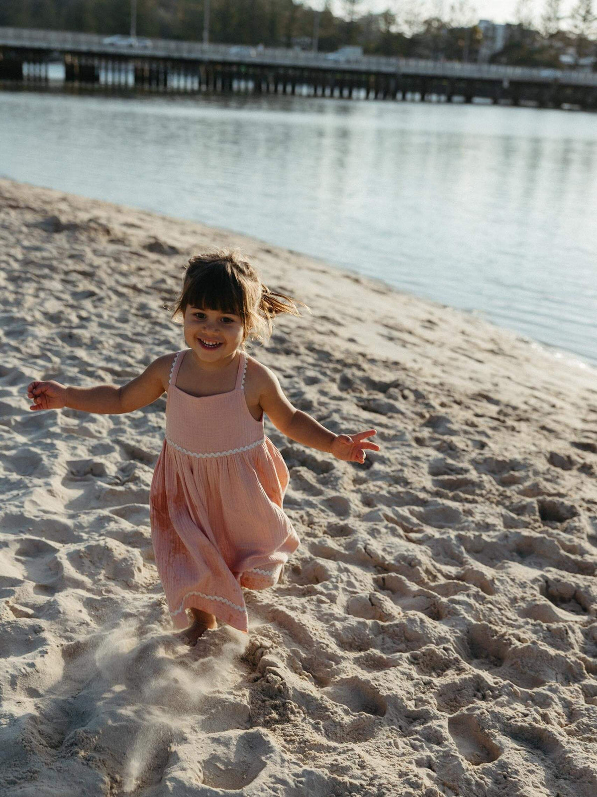A young girl smiling and running on a sandy beach next to the water, wearing a light pink sleeveless dress.
