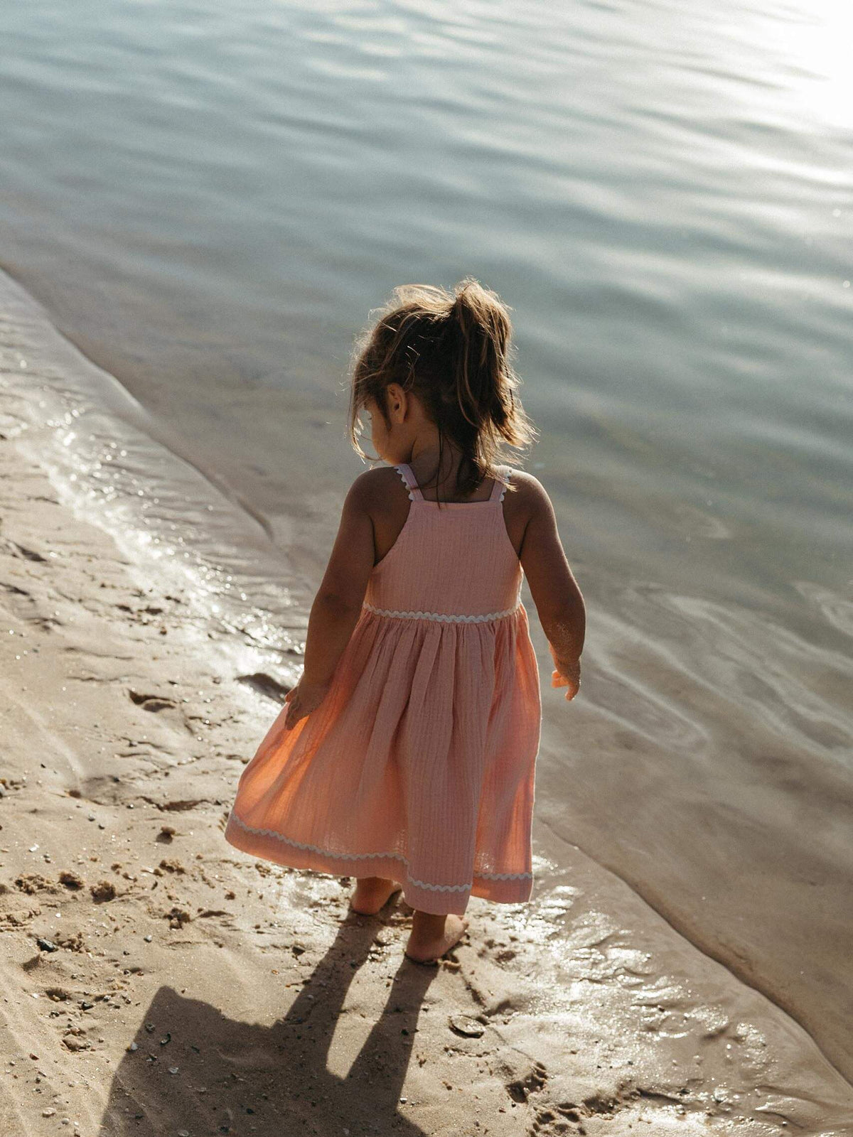 A young girl with her hair in a ponytail stands on a sandy beach at the water's edge, wearing a sleeveless dusty pink dress with white trim.
