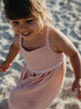 A young child wearing a light pink textured sleeveless dress with white scalloped trim, standing on a sandy beach.