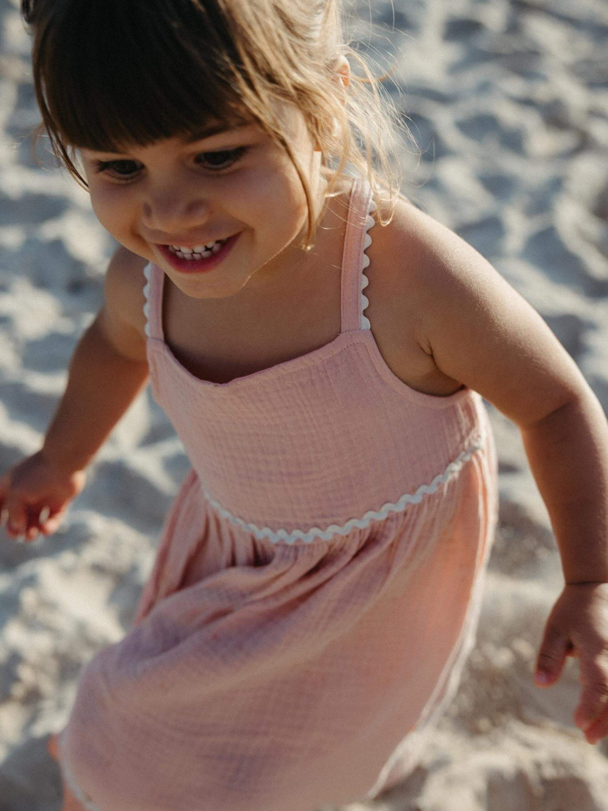 A young child wearing a light pink textured sleeveless dress with white scalloped trim, standing on a sandy beach.