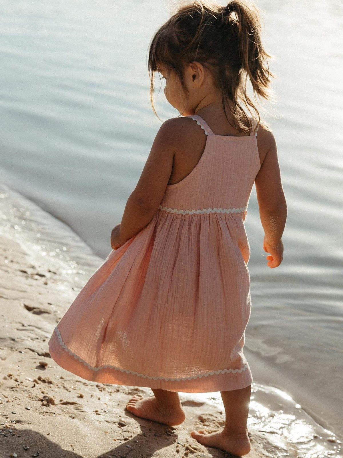 A toddler girl stands barefoot on a sandy beach at the water's edge, wearing a sleeveless light pink dress with white scalloped trim.