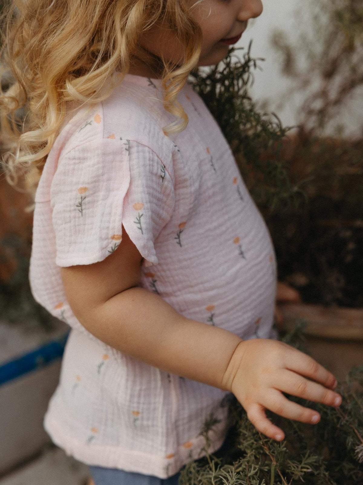 A young child wearing a light pink gauze short-sleeve t-shirt with a small floral print, standing outdoors next to some green plants.