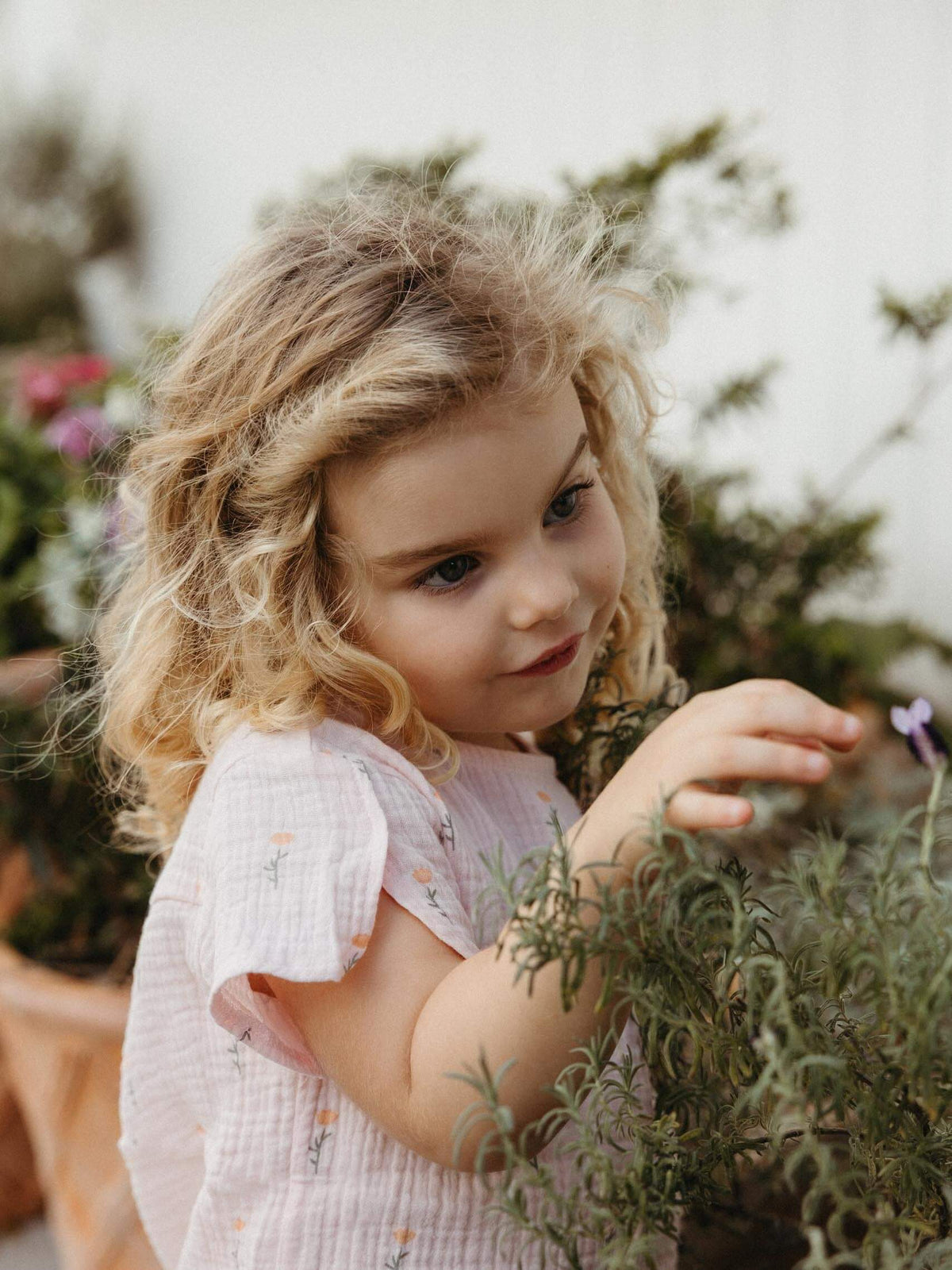 A young girl with curly blonde hair wearing a light pink short-sleeve dress, looking down at green plants in a garden setting.