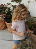 A young girl with curly blonde hair wearing a light pink textured short-sleeve top with a tiny floral print and blue denim shorts, standing in a garden next to potted plants.