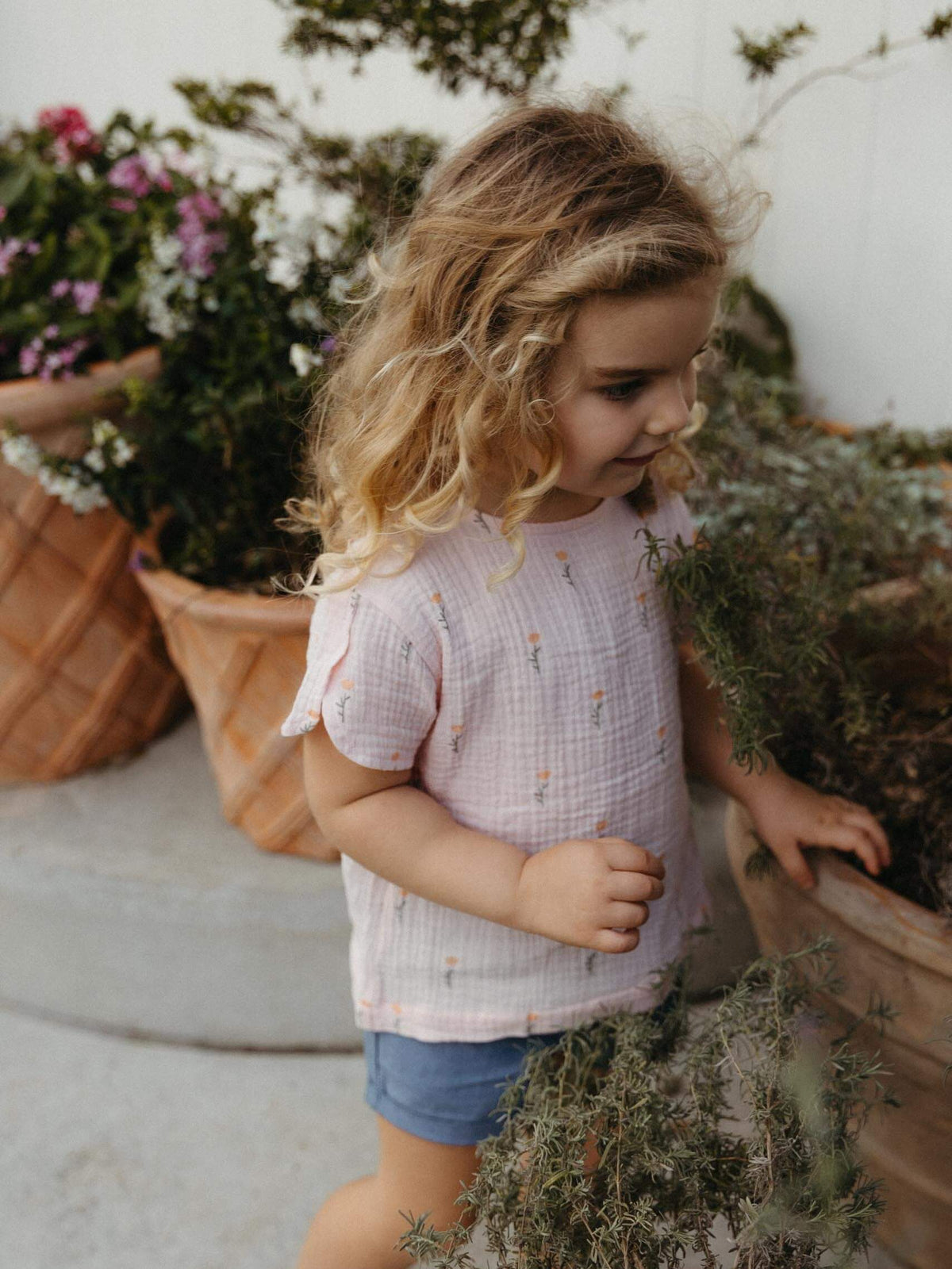 A young girl with curly blonde hair wearing a light pink textured short-sleeve top with a tiny floral print and blue denim shorts, standing in a garden next to potted plants.