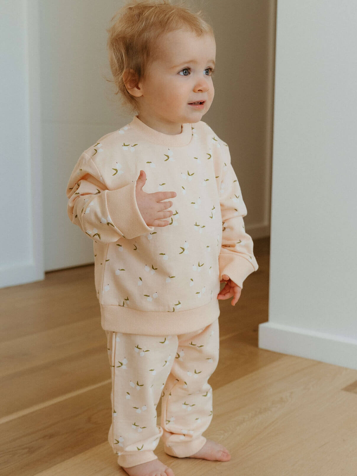 Child wearing a light pink outfit with small patterns indoors on a wooden floor.