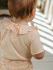 Baby wearing a light-colored outfit with a ruffled collar, standing in a softly lit room.