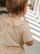 Baby wearing a light-colored outfit with a ruffled collar, standing in a softly lit room.
