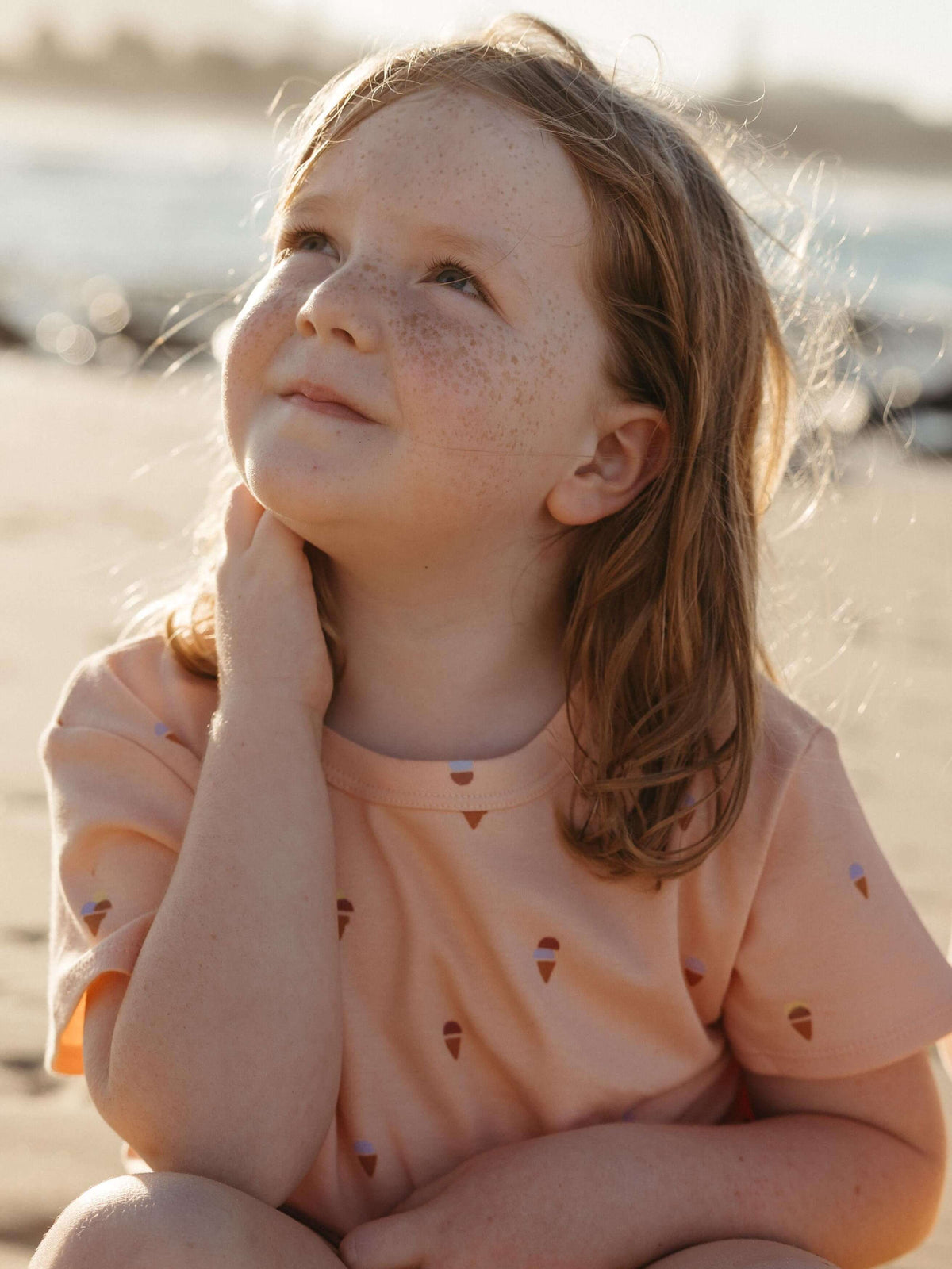 A young girl with freckles sits on a sandy beach, looking upward and to the side. She is wearing a peach-colored short-sleeve t-shirt with a small repeating pattern of ice cream cones.