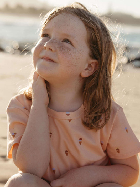 A young girl with freckles sits on a sandy beach, looking upward and to the side. She is wearing a peach-colored short-sleeve t-shirt with a small repeating pattern of ice cream cones.