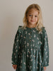 Young girl wearing a green dress with a pattern of small figures against a plain background Printed Woven Dress - Forest Gathering Balsam Girls