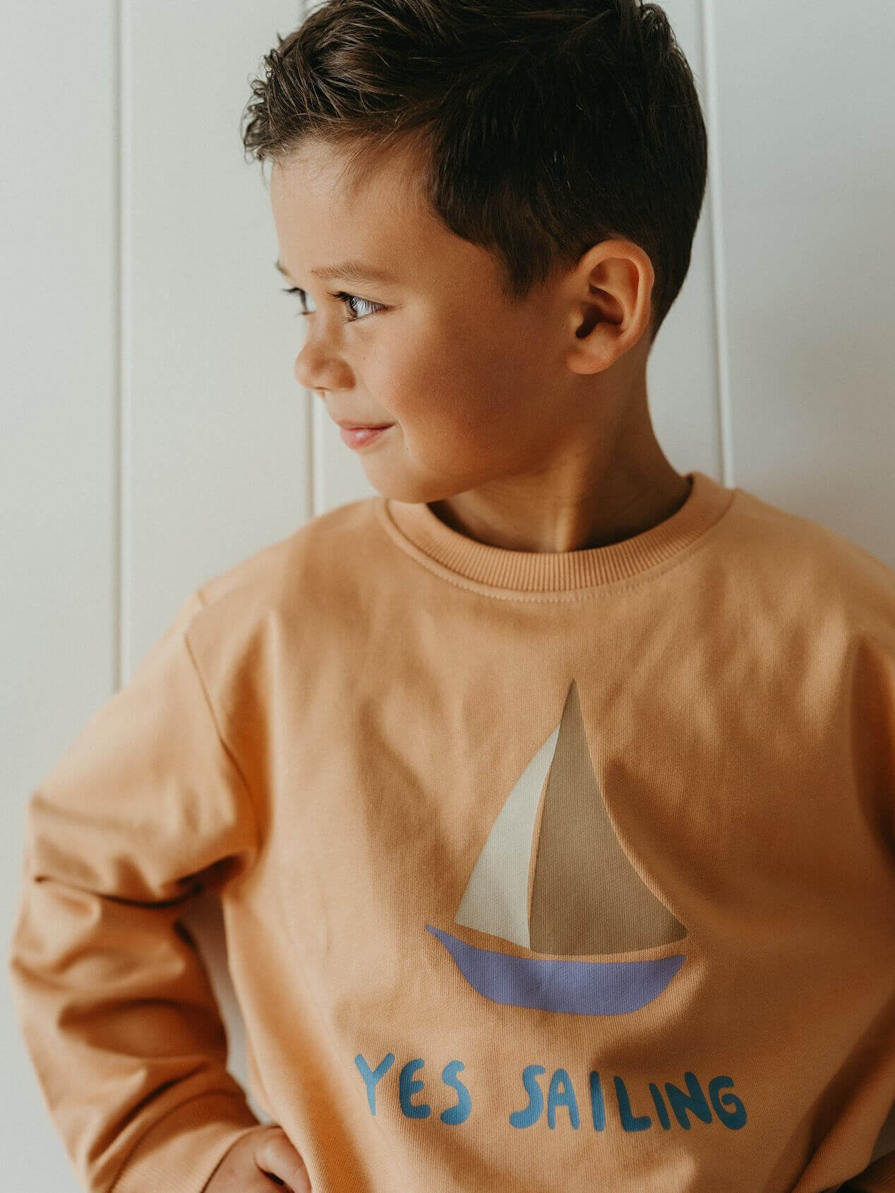 Child wearing a brown sweatshirt with a sailboat design and 'YES SAILING' text.