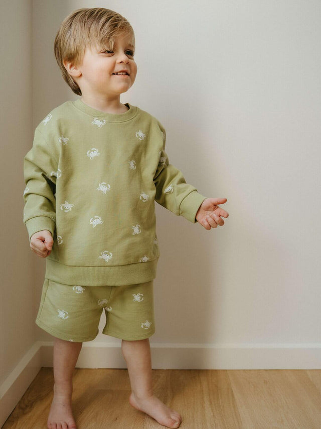 Child wearing a green outfit with white patterns standing on a wooden floor.