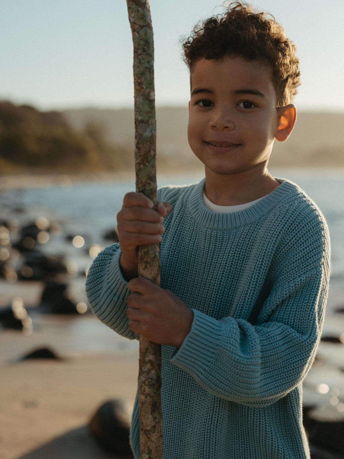 Boy holding a stick at the beach wearing a light blue knit sweater