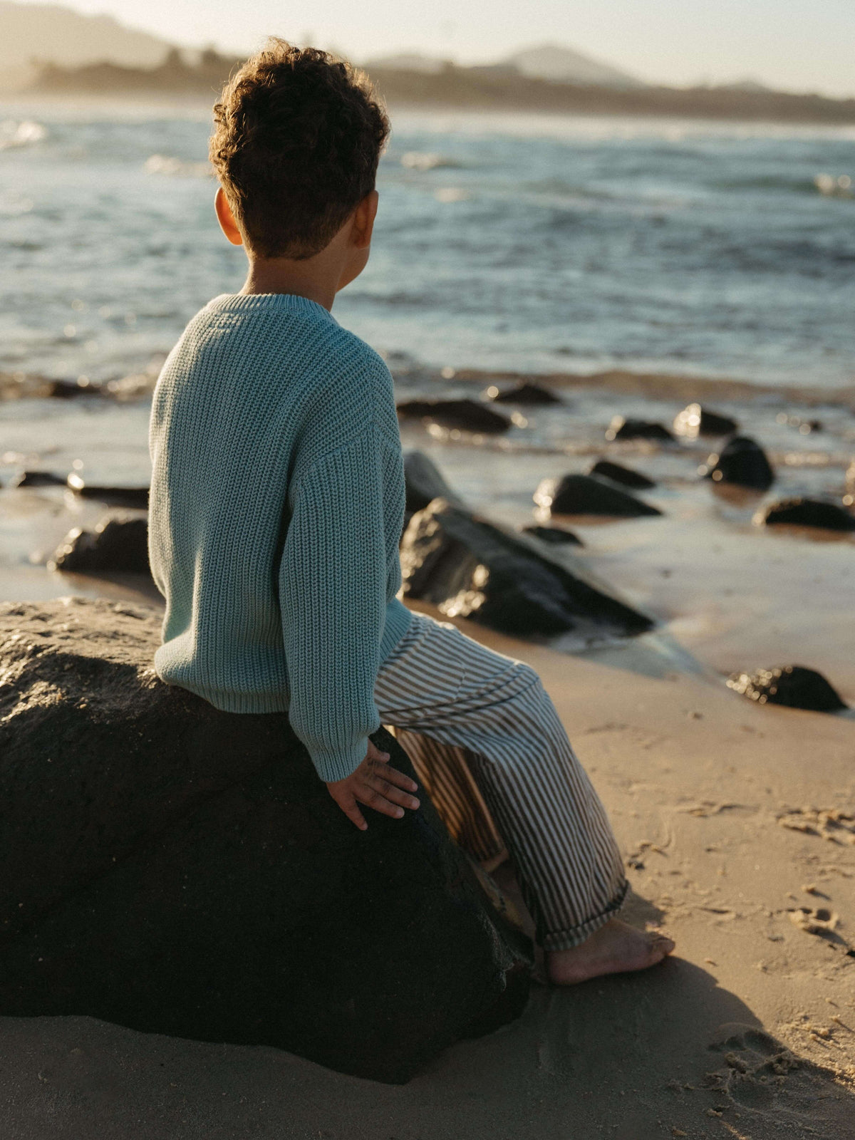 Boy looking at the sea on a beautiful sunset at the beach wearing a blue ribbed knit jumper