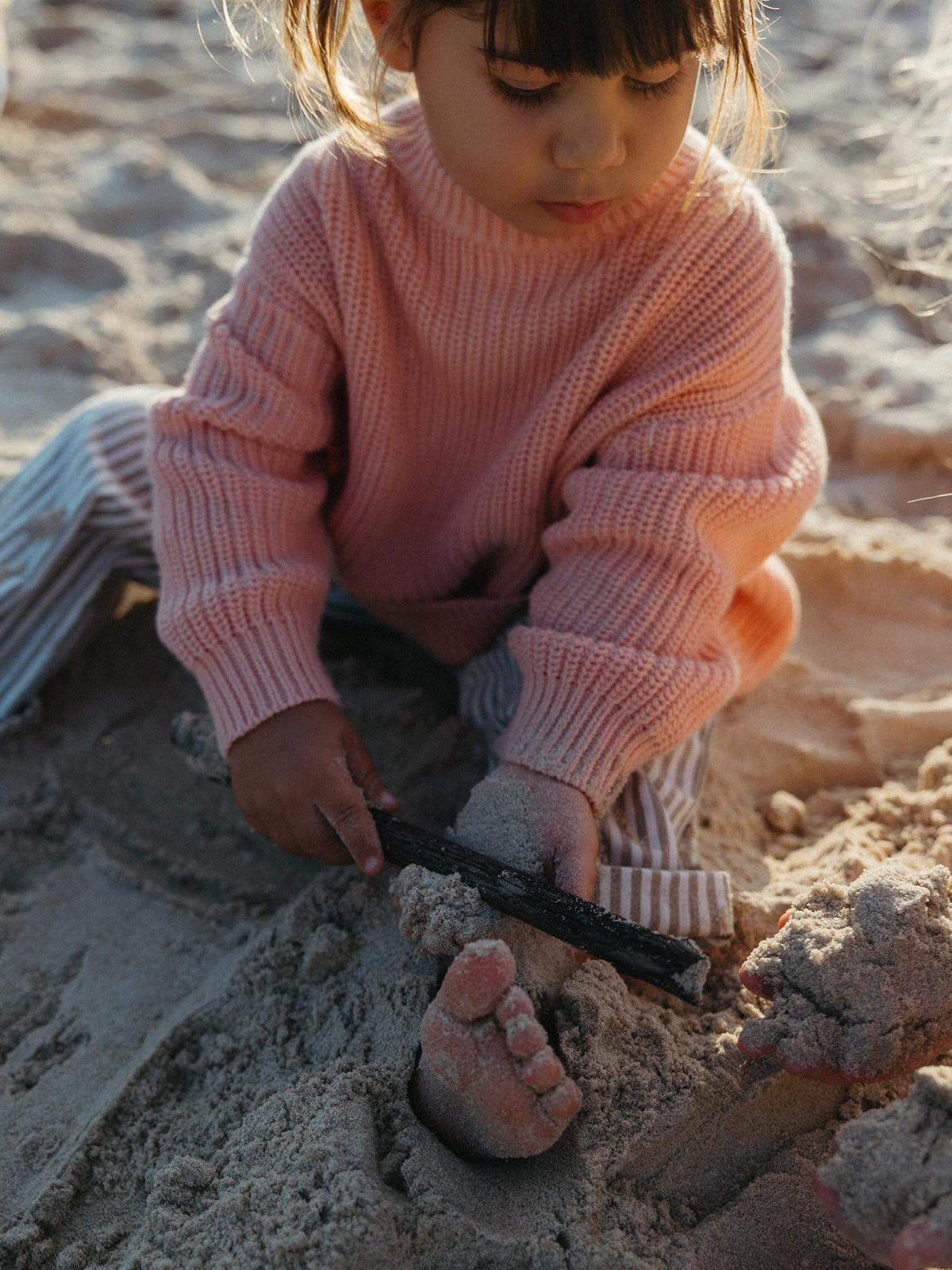 A young child wearing a pink ribbed knit sweater and striped pants sits in the sand, using a small black tool to dig or shape sand near their bare foot.