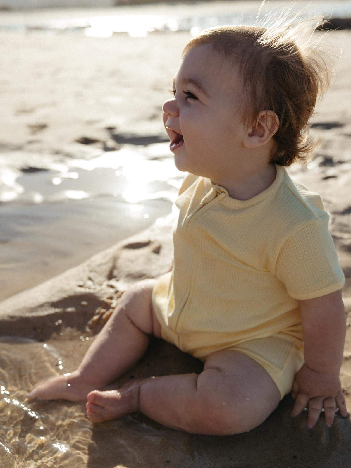 A baby sitting on wet sand at the beach wearing a pale yellow ribbed short-sleeve bodysuit.