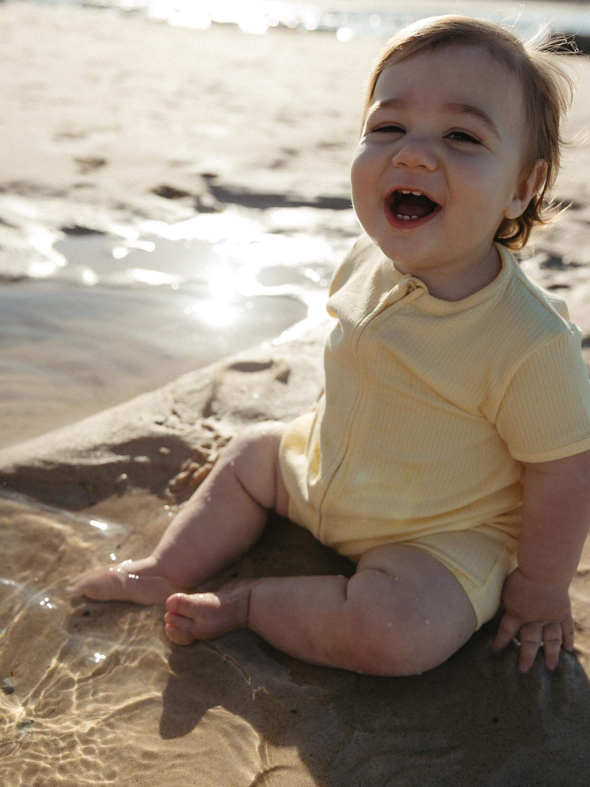 A baby sitting on wet sand at the beach wearing a pale yellow short-sleeve romper, looking up and smiling with the sun reflecting on the water behind them.