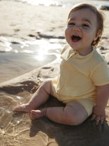 A baby sitting on wet sand at the beach wearing a pale yellow short-sleeve romper, looking up and smiling with the sun reflecting on the water behind them.