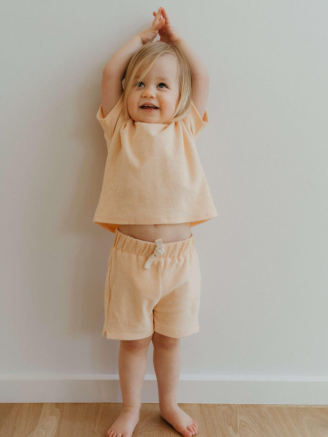 Young child in peach outfit with arms raised against white wall Riley Towelling Shorts - Apricot Baby girl