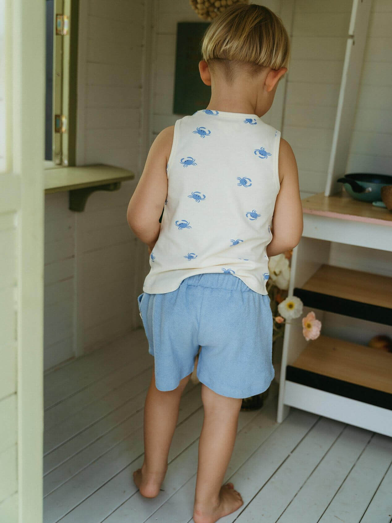 Child wearing a sleeveless shirt with blue patterns and light blue shorts in a room with wooden floors and white walls.