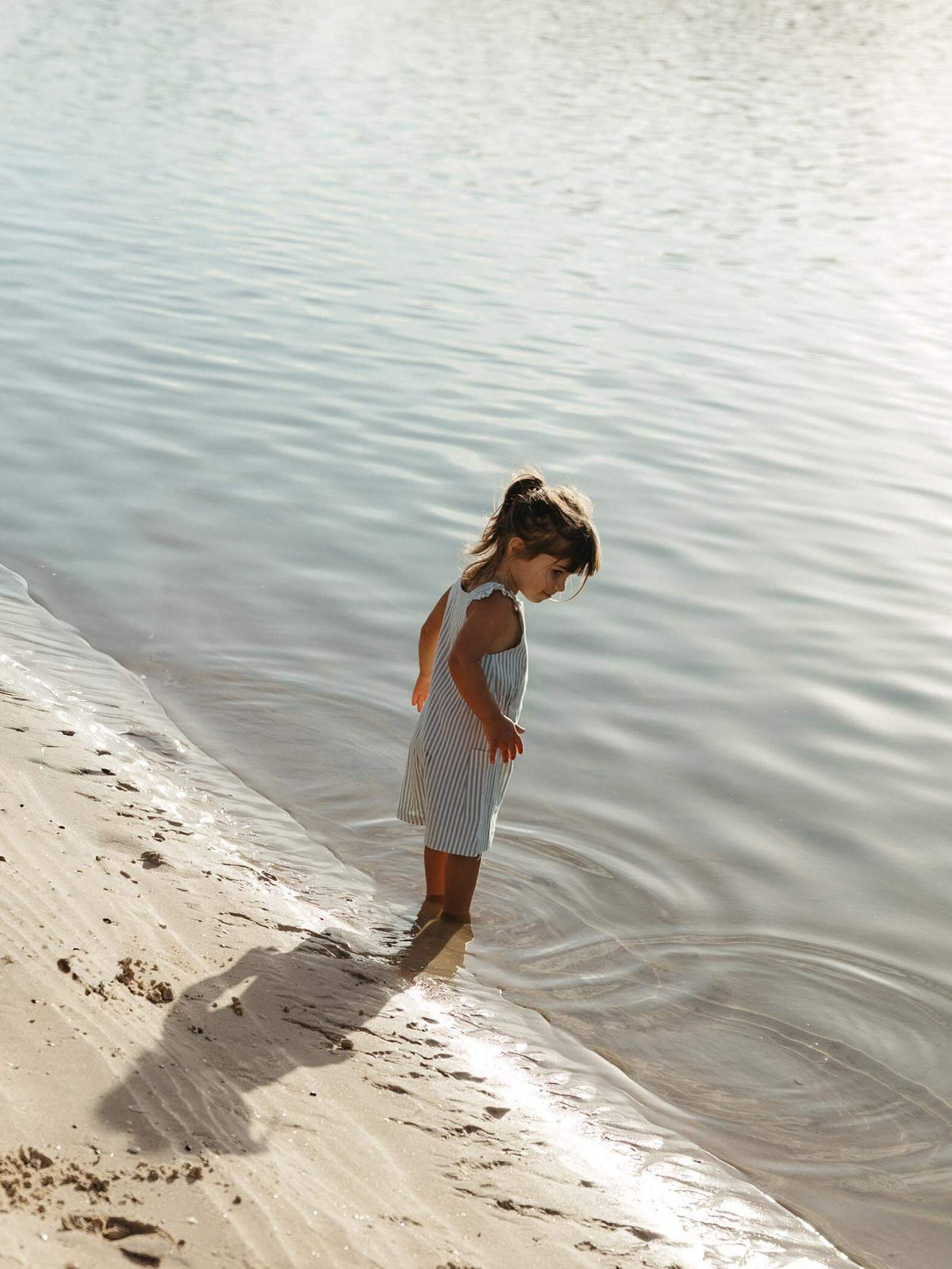 A young child stands in shallow water at the shoreline, wearing a sleeveless light blue and white vertically striped dress. The background is a natural outdoor setting with calm water and wet sand.