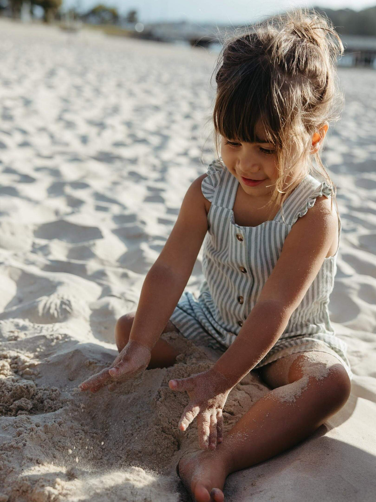 A young girl with her hair in a messy bun sits on a sandy beach, playing with sand while wearing a sleeveless, light blue and white striped romper with gold buttons.